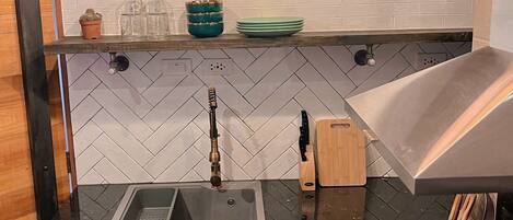 A kitchen with a black granite countertop, a stainless steel sink, a wooden shelf with glassware, and a modern stove.