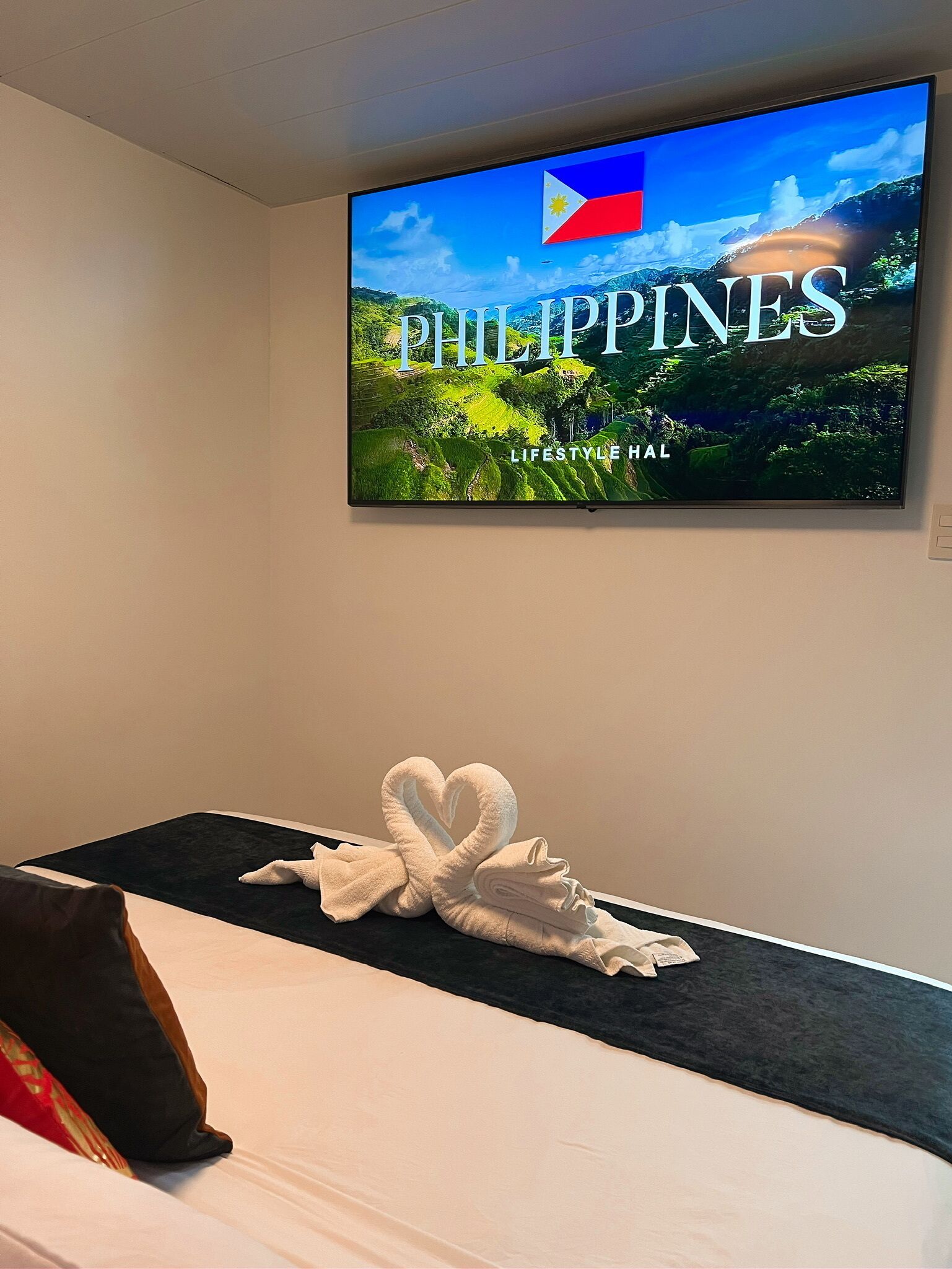 A hotel room with a bed, a wall-mounted TV displaying 'PHILIPPINES LIFESTYLE HAL', and a towel art on the bed.