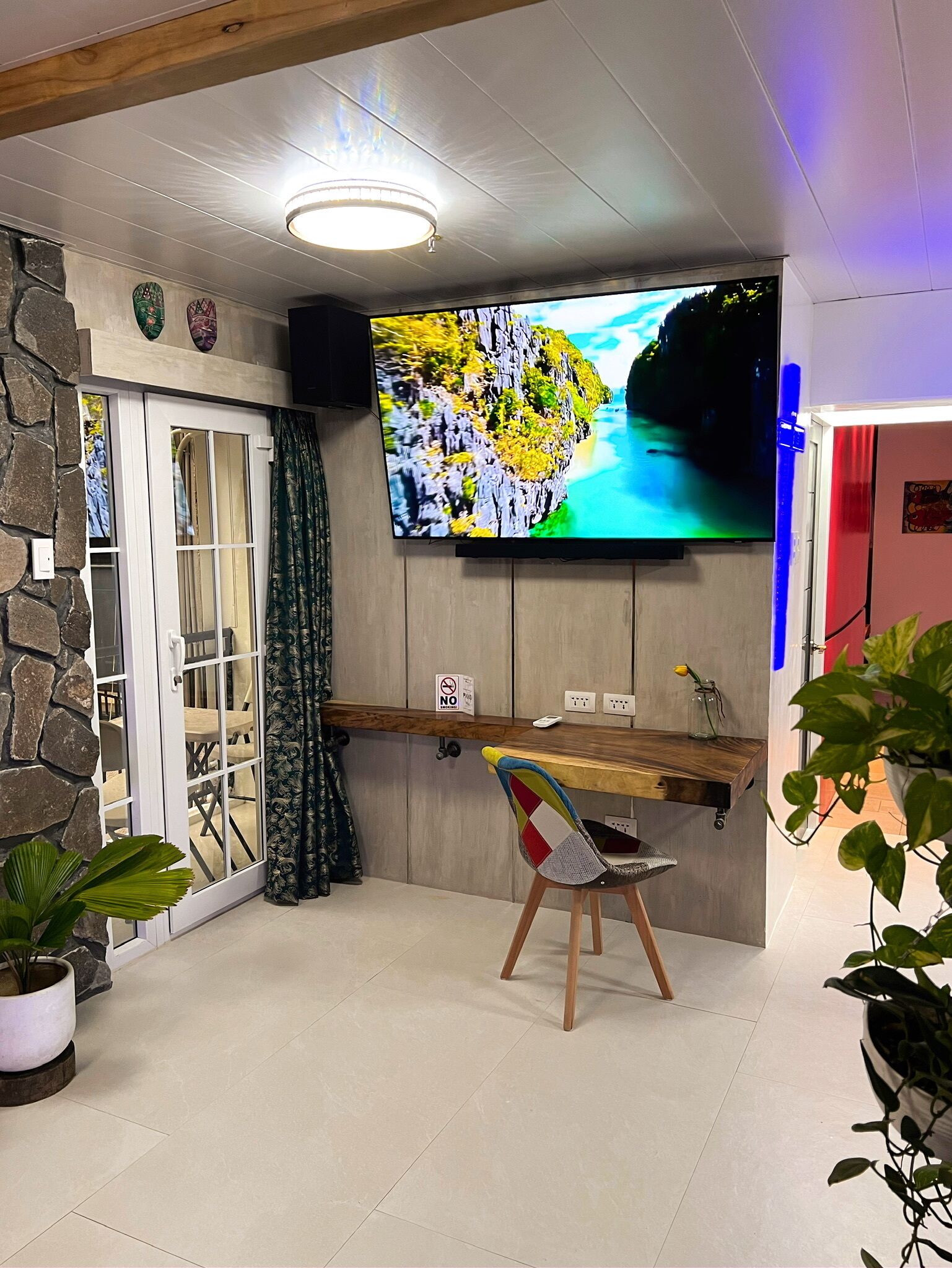 Een kamer met een stenen muur, een houten bureau, een aan de muur bevestigde televisie, een stoel en potplanten.