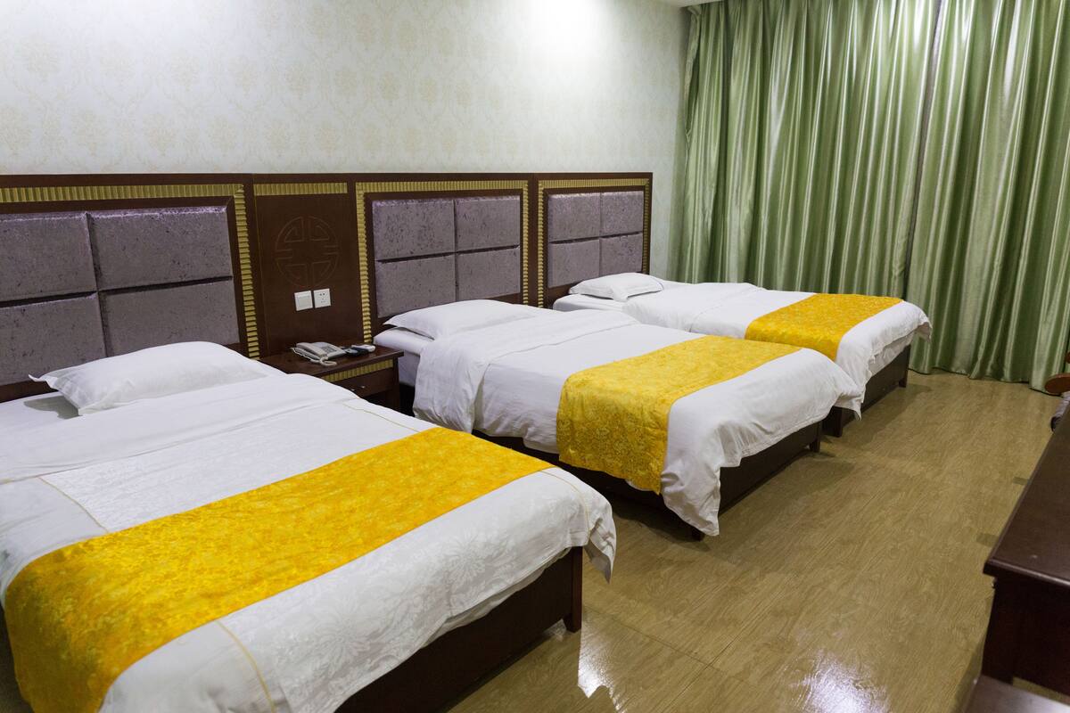 A hotel room with three beds, each with a white bedspread and a yellow accent. The room has a wooden headboard, a wooden nightstand, and a green curtain.