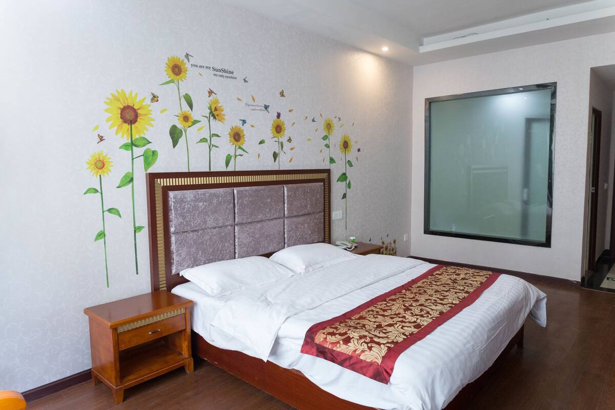 A neatly made bed with a patterned bedspread, a wooden nightstand with a drawer, and a wall with a floral mural.