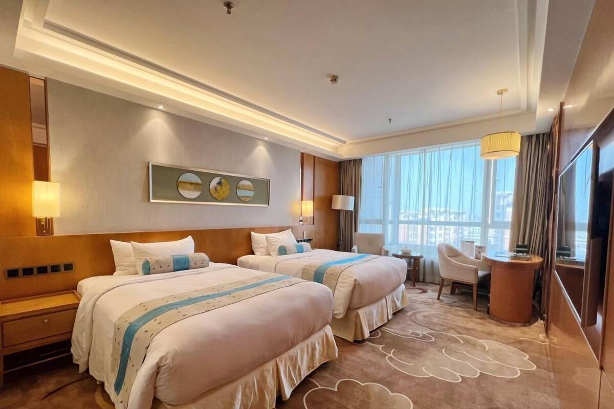 A hotel room with two beds, a sitting area with a chair, a small table, and a large window with curtains.