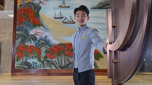 A man in a uniform standing in front of a large mural depicting a landscape with water, boats, and a bridge. - Menshine Gloria Plaza Hotel Shantou (Shantou)