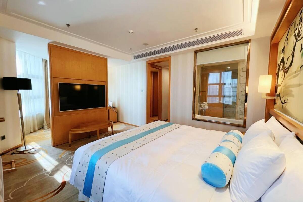A hotel room with a large bed, a flat-screen TV mounted on the wall, a desk, and a large window.