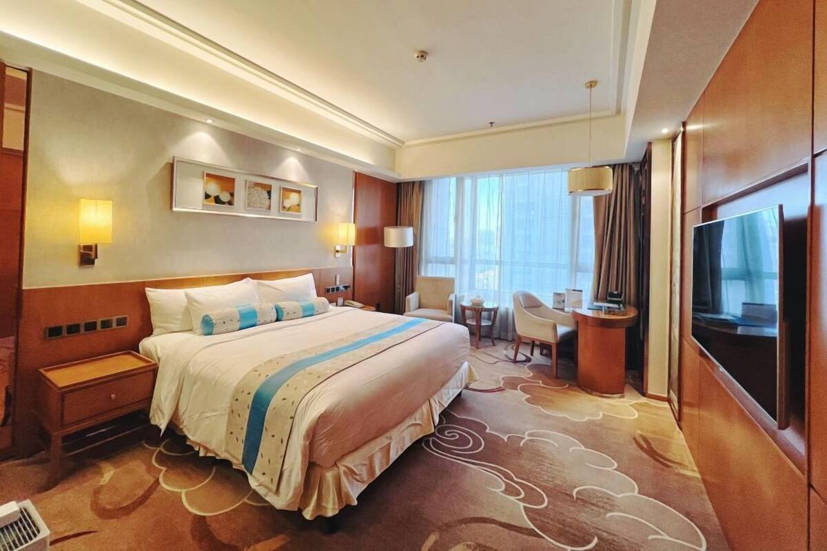 A hotel room with a large bed, a desk, a chair, a TV, and a window with curtains.