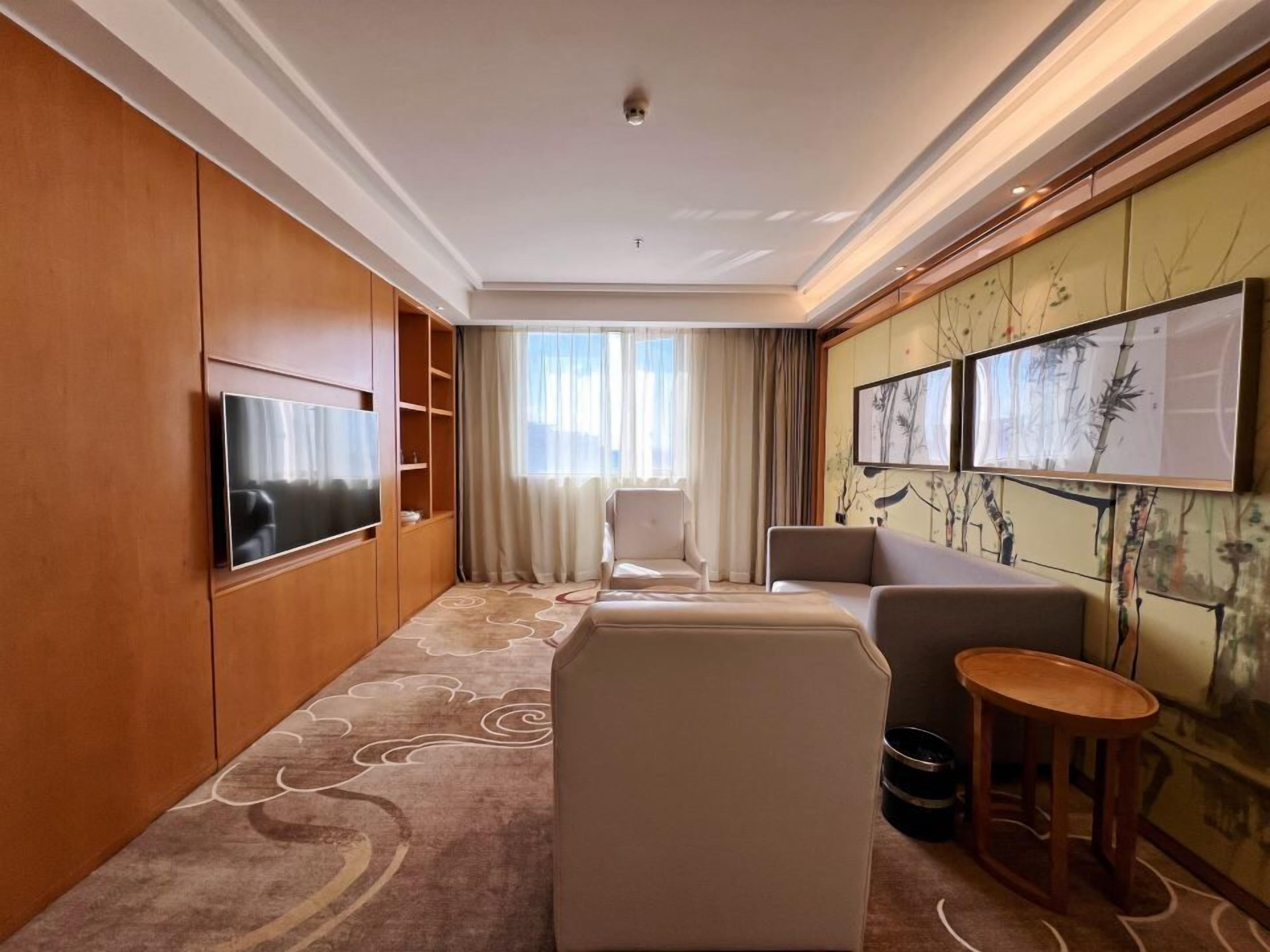 A hotel room with a flat-screen TV, a sofa, a chair, a small wooden table, and artwork on the wall.