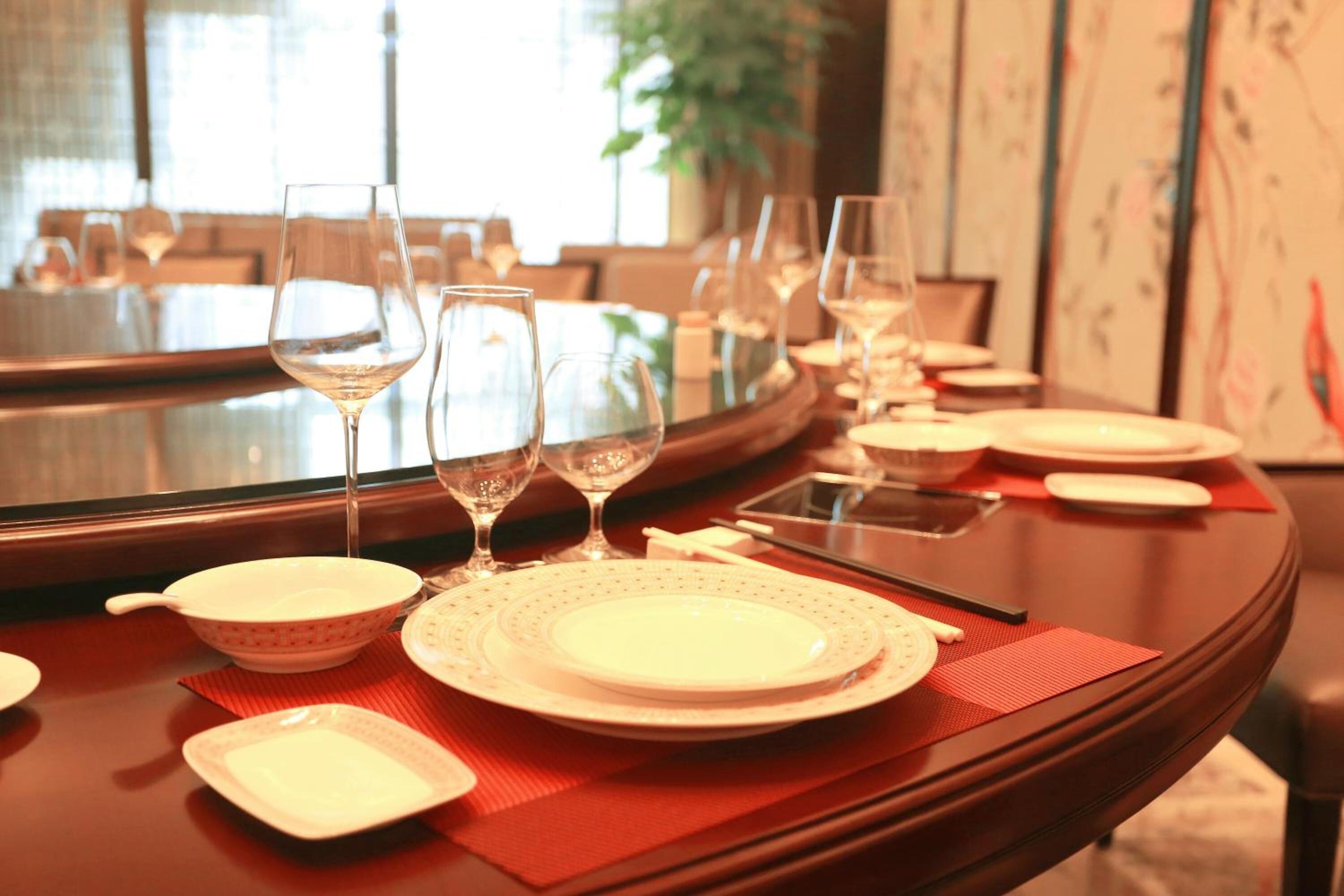 Una mesa de comedor con platos, tazones y copas de vino blancos, un camino de mesa rojo y palillos.