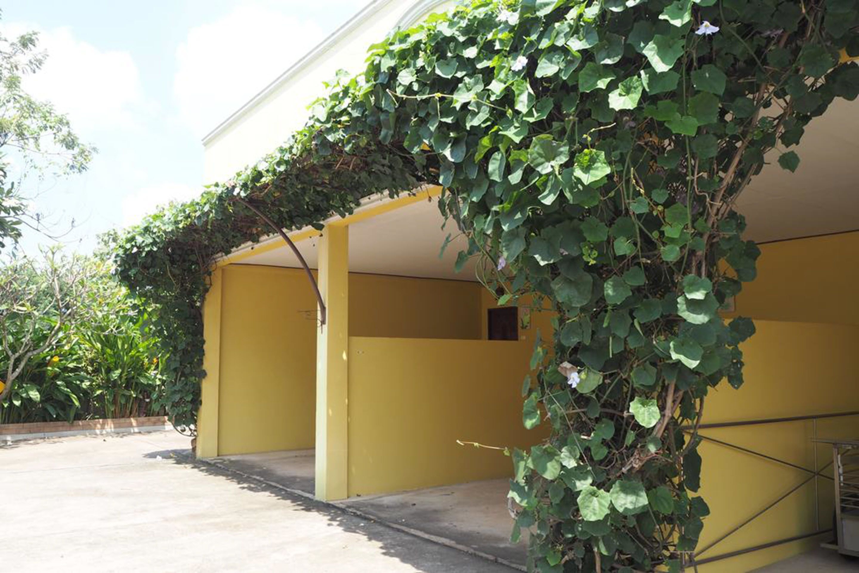 A yellow building with a covered outdoor area, surrounded by greenery.
