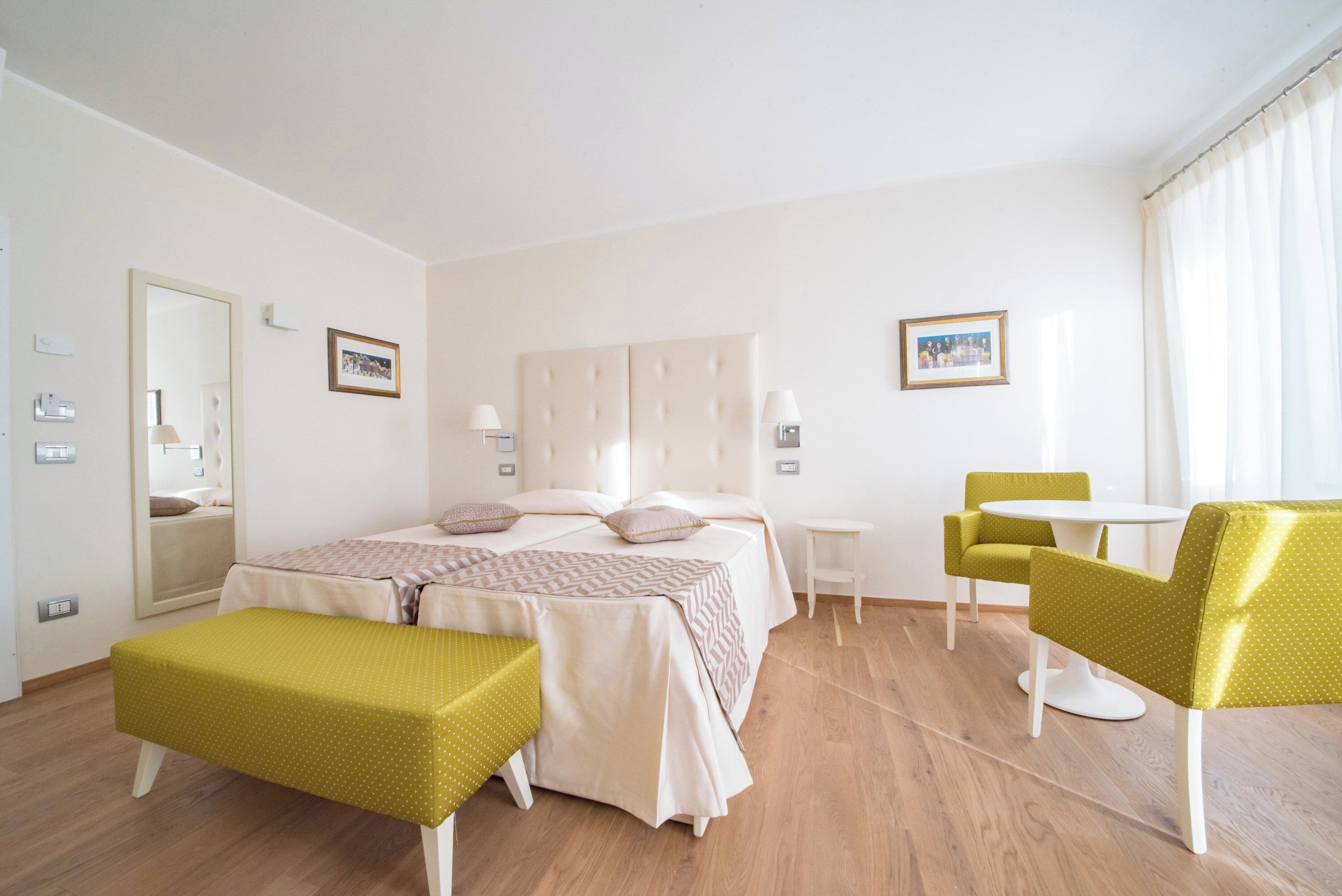 A hotel room with a bed, a small table, two chairs, a bench, and a mirror.