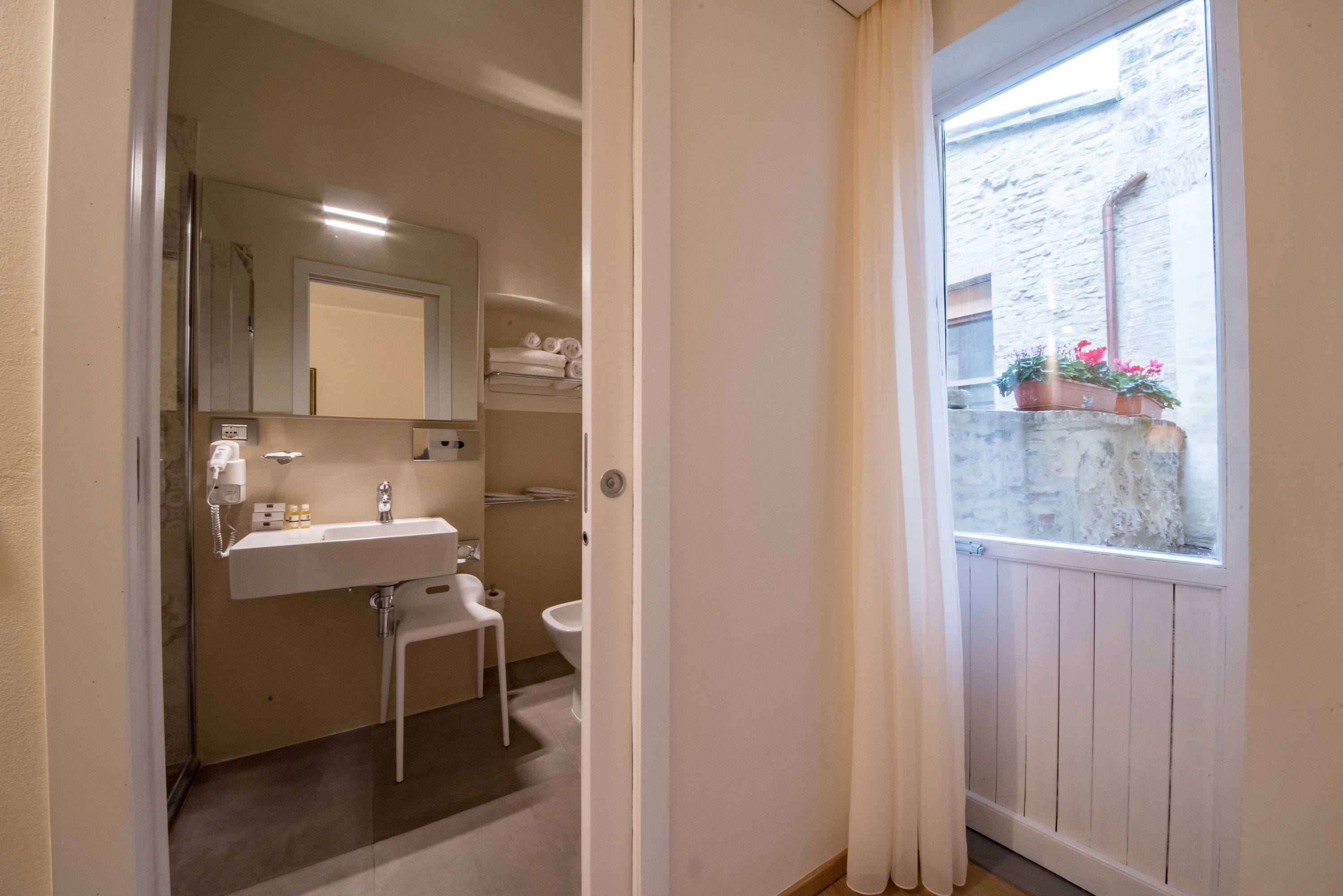 A bathroom with a sink, mirror, and toilet. There is a window with curtains and a view of a stone wall outside.