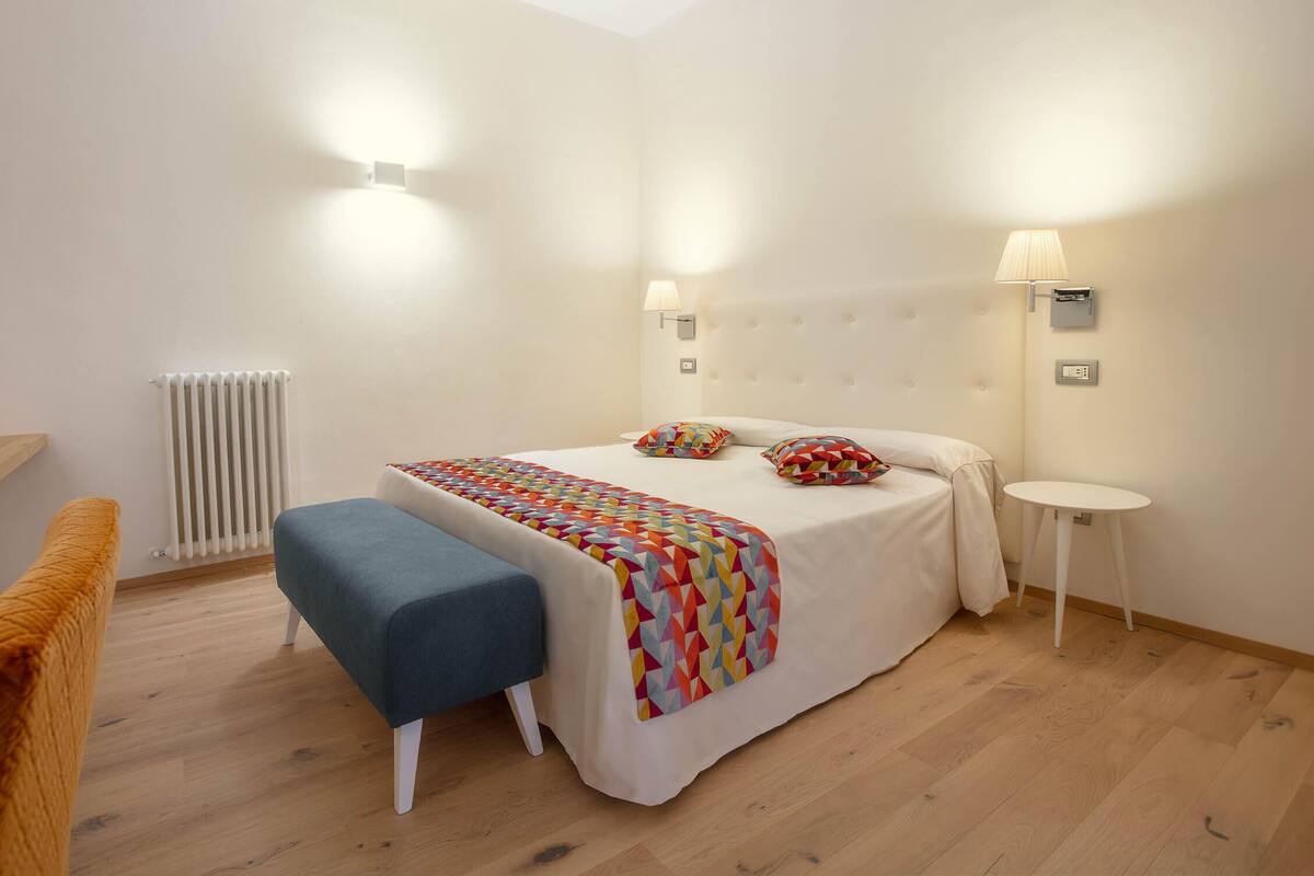 Un dormitorio con cama, lámparas de noche, un radiador, un banco y una mesita.