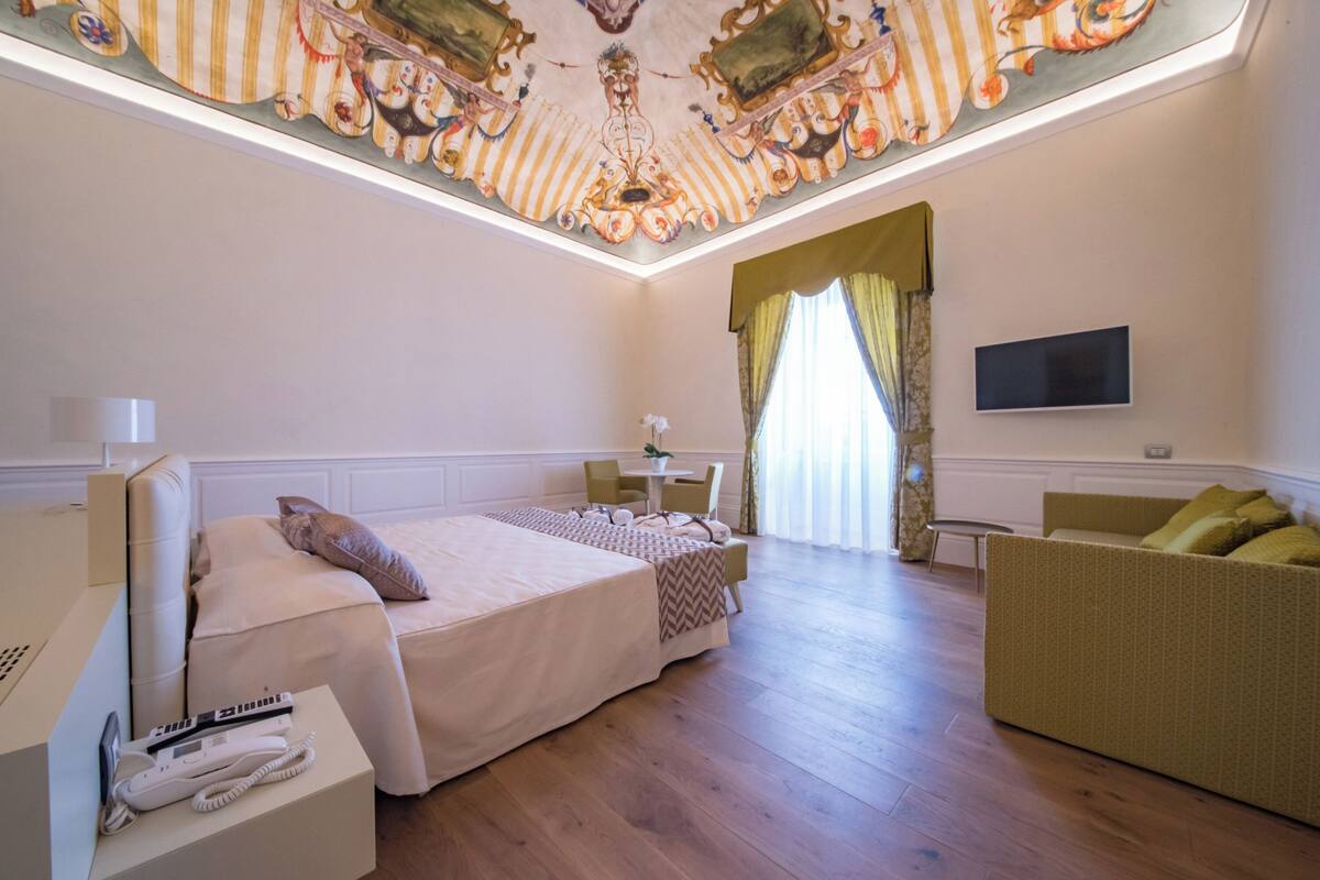 Una habitación de hotel con una cama grande, un sofá, un televisor y un techo con detalles artísticos.