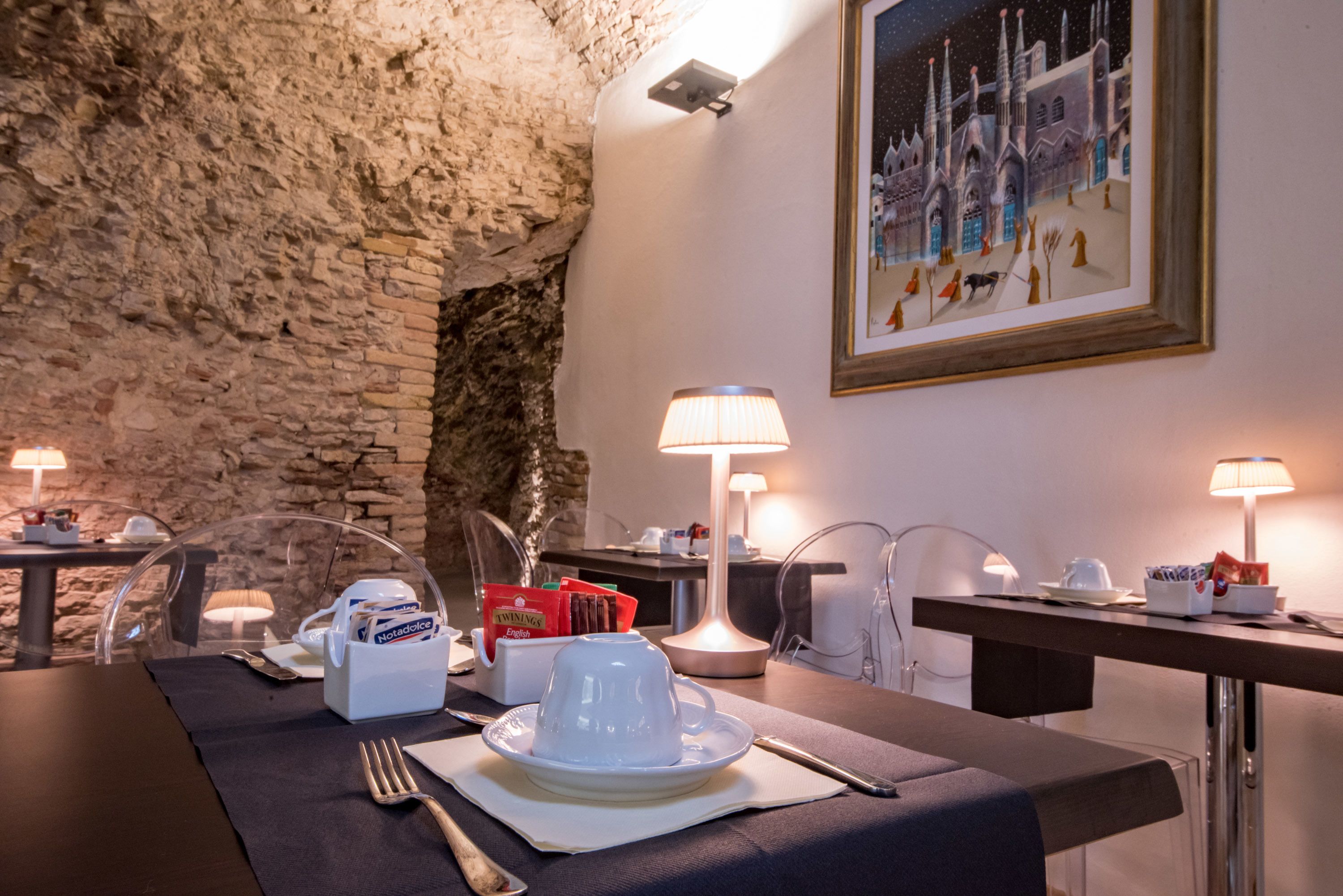 A dining area with a stone wall, a table set with a white plate, a cup, and a Notadolce coffee machine. There are two lamps on the table and a framed picture on the wall.
