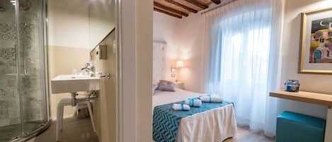 A hotel room with a shower, a sink, a bed with a blue and white bedspread, a bedside table with a lamp, and a painting on the wall.