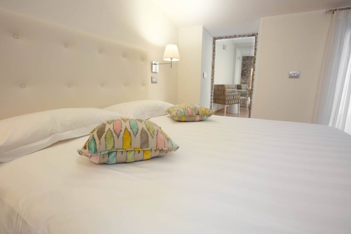 Un dormitorio con una cama grande, dos almohadillas decorativas, una lámpara fijada a la pared y vistas a un baño por una puerta abierta.