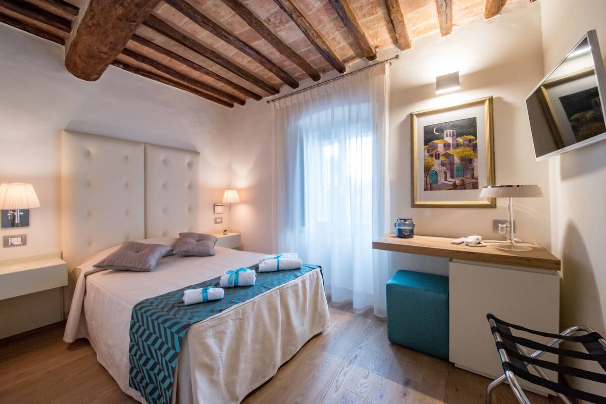 Una habitación de hotel con una cama, mesitas de noche, un escritorio, una silla, un archivador pequeño y un cuadro en la pared.