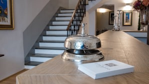 Un comptoir d’accueil en bois, agrémenté d’une clochette argentée et d’une pile de cartes blanches. Un escalier avec une rampe en métal est visible au fond.
