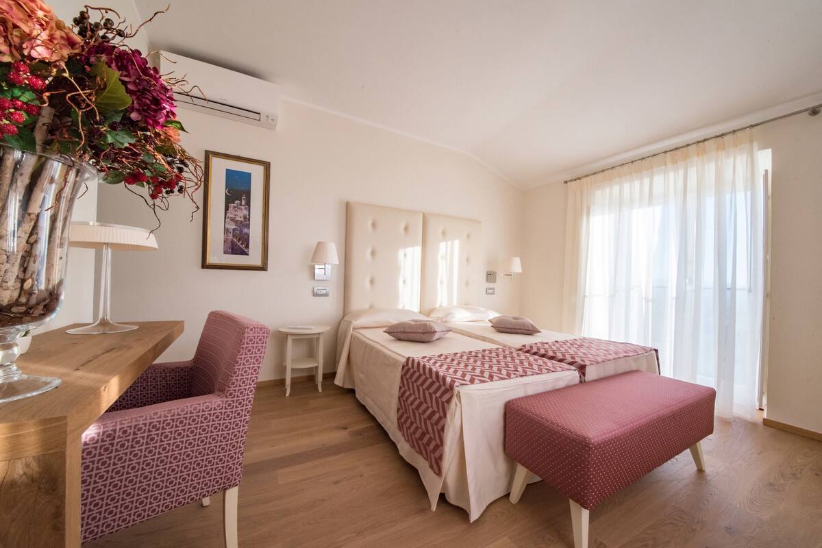Un dormitorio con cama, mesitas de noche, una silla, un escritorio, un jarrón con flores y una ventana con cortinas.