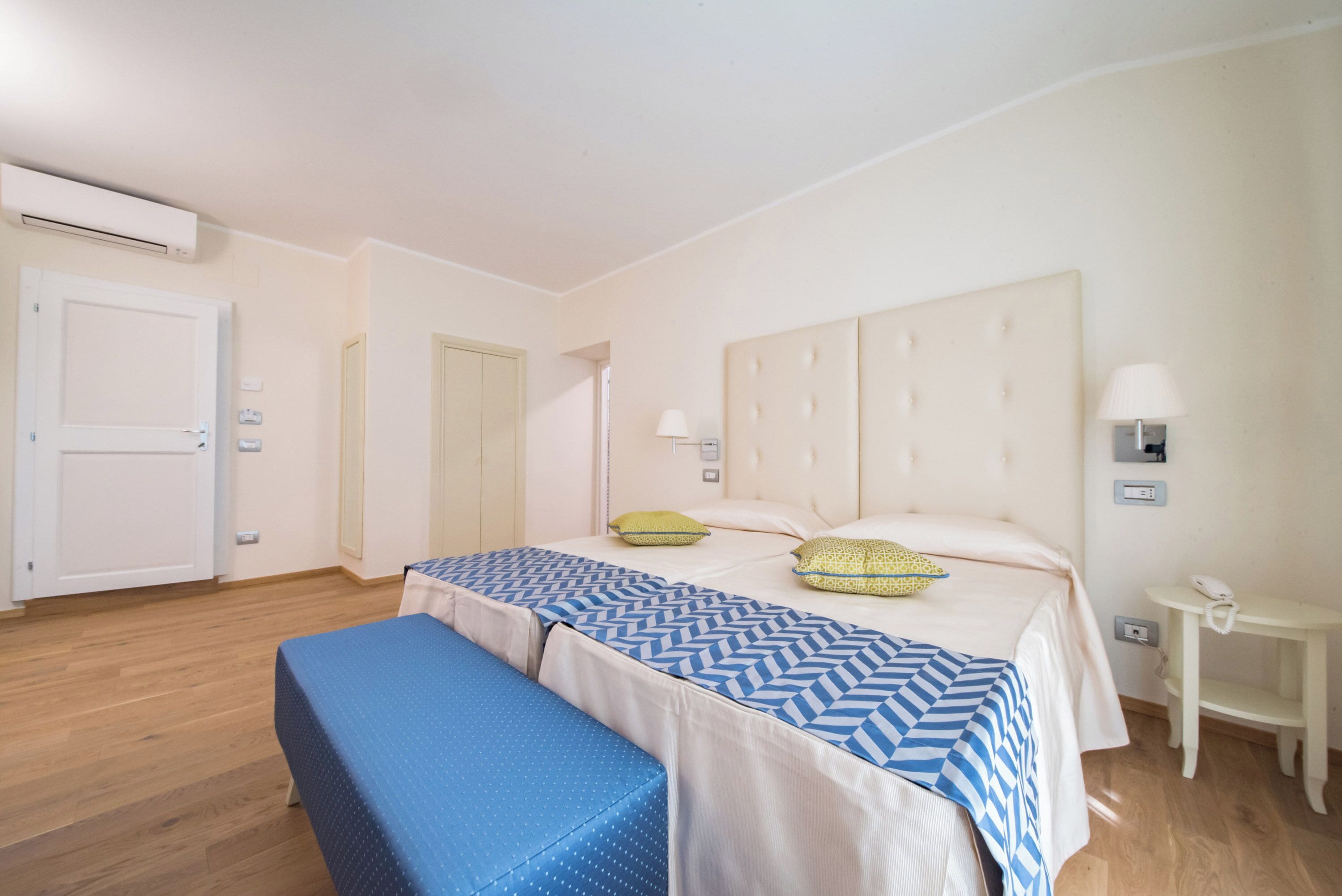 A bedroom with two beds, a bench, a bedside table, and an air conditioning unit.