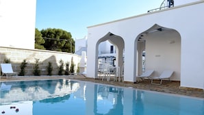 A swimming pool with lounge chairs and a table, surrounded by white walls and stone flooring.