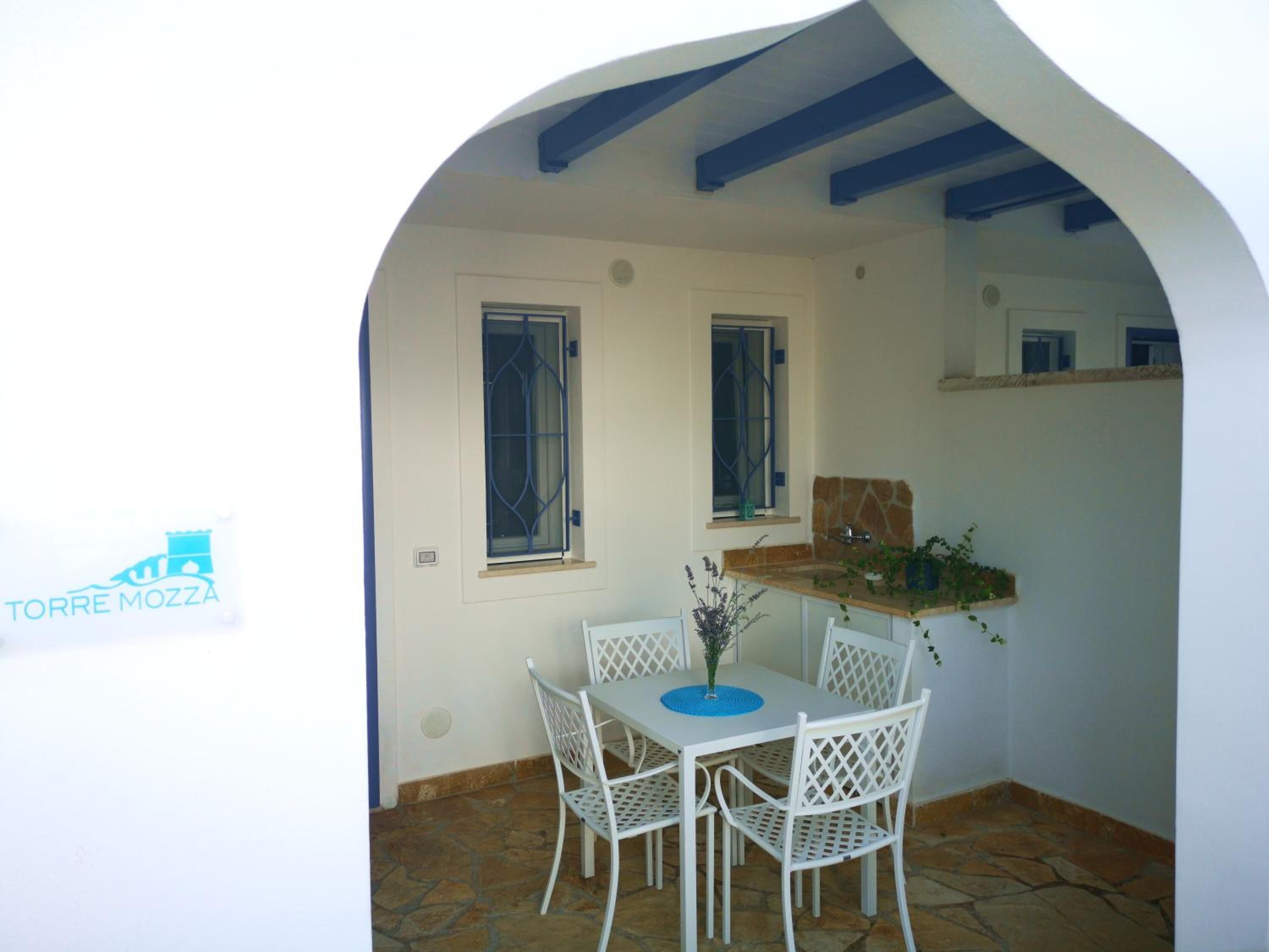 A small outdoor dining area with a white table and chairs, a stone floor, and a blue railing.