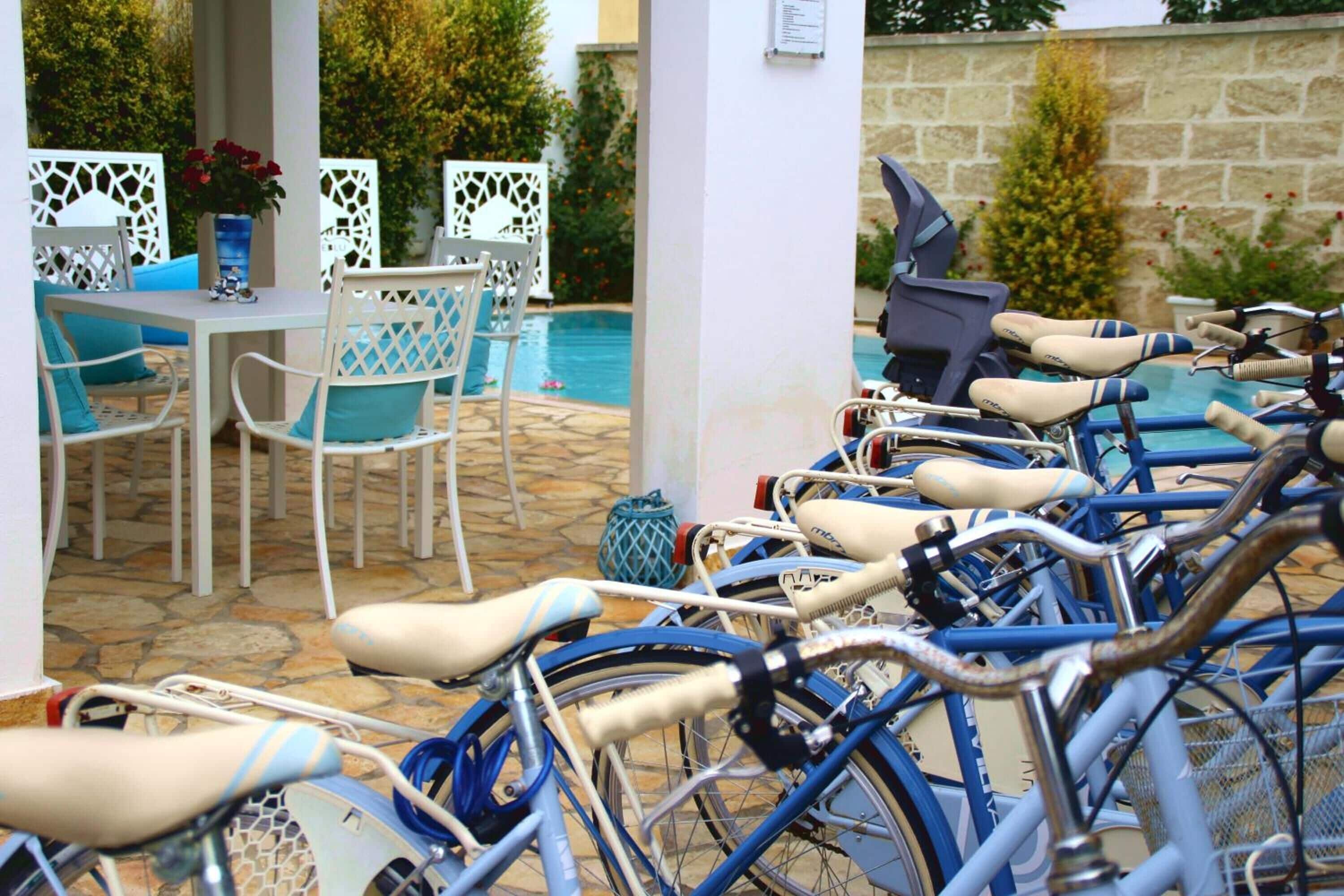 Varias bicicletas aparcadas cerca de una piscina, con sillas y una mesa blancas.