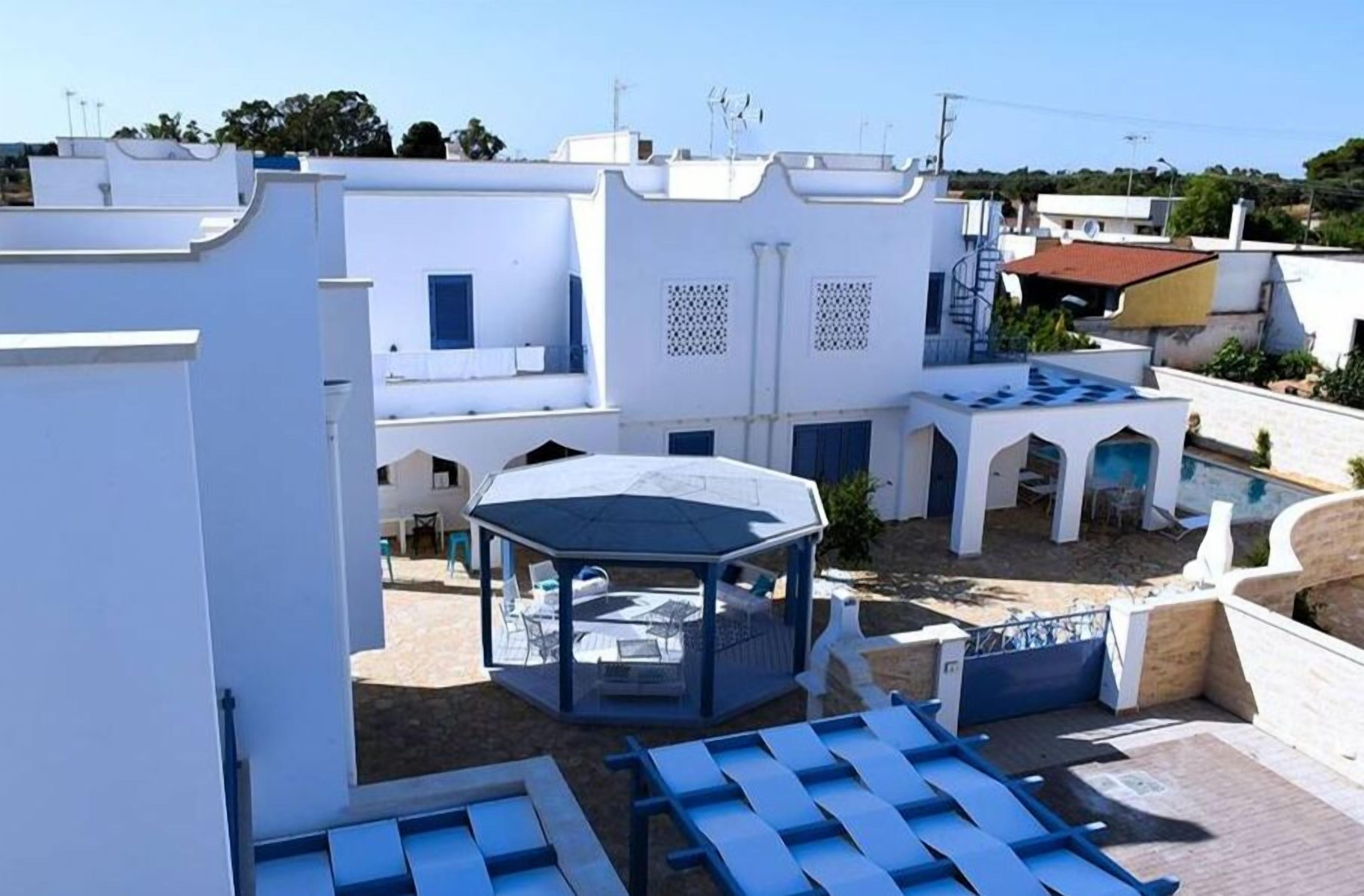 Un cortile con un gazebo esagonale, edifici bianchi e poltrone da relax blu.