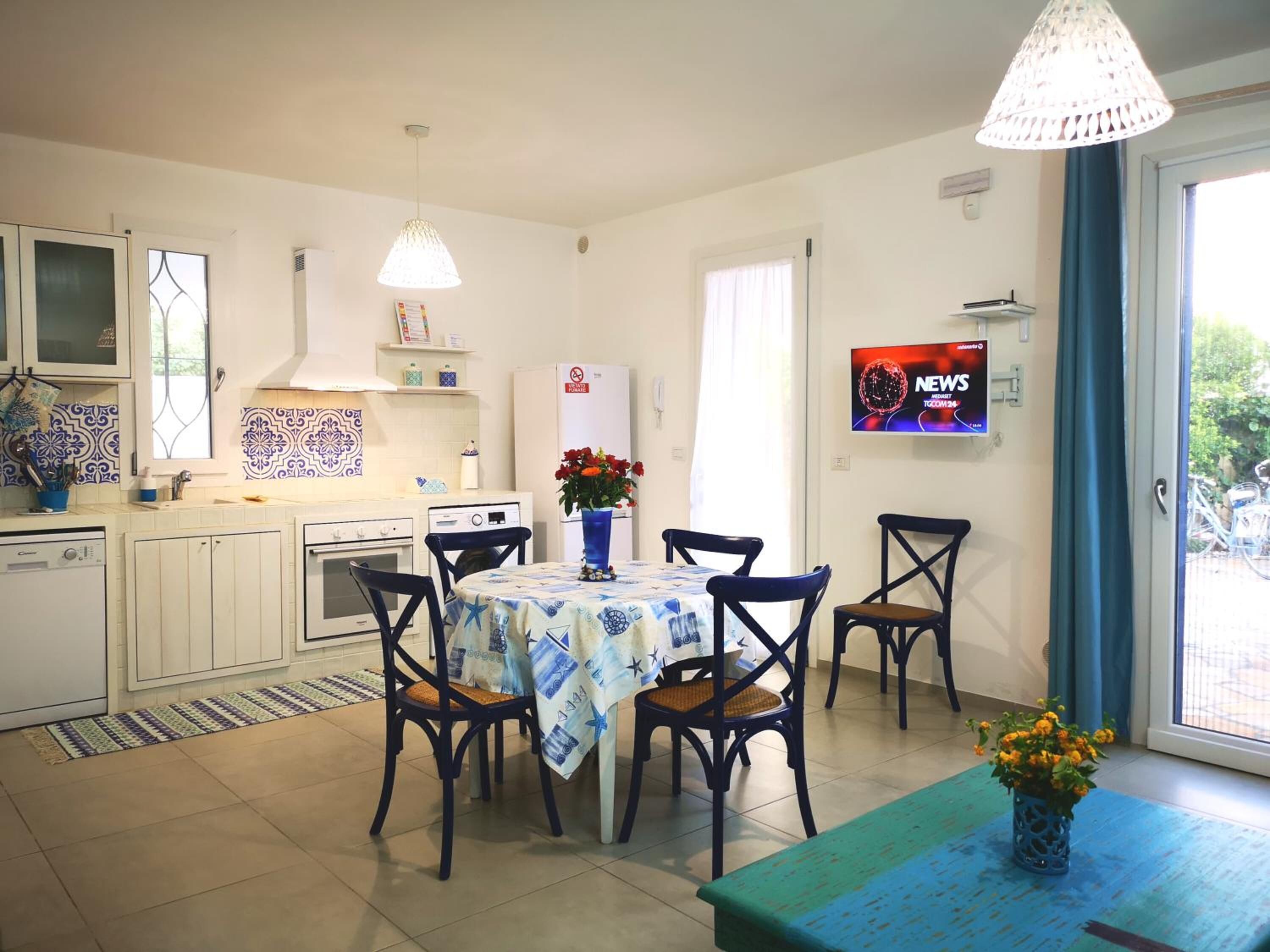 Eine kleine Küche mit weißen Schränken, einem Esstisch mit blauer Tischdecke und einem Fernseher an der Wand.
