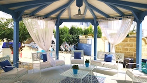 A covered outdoor seating area with white furniture and blue accents, featuring a patterned rug and potted plants.