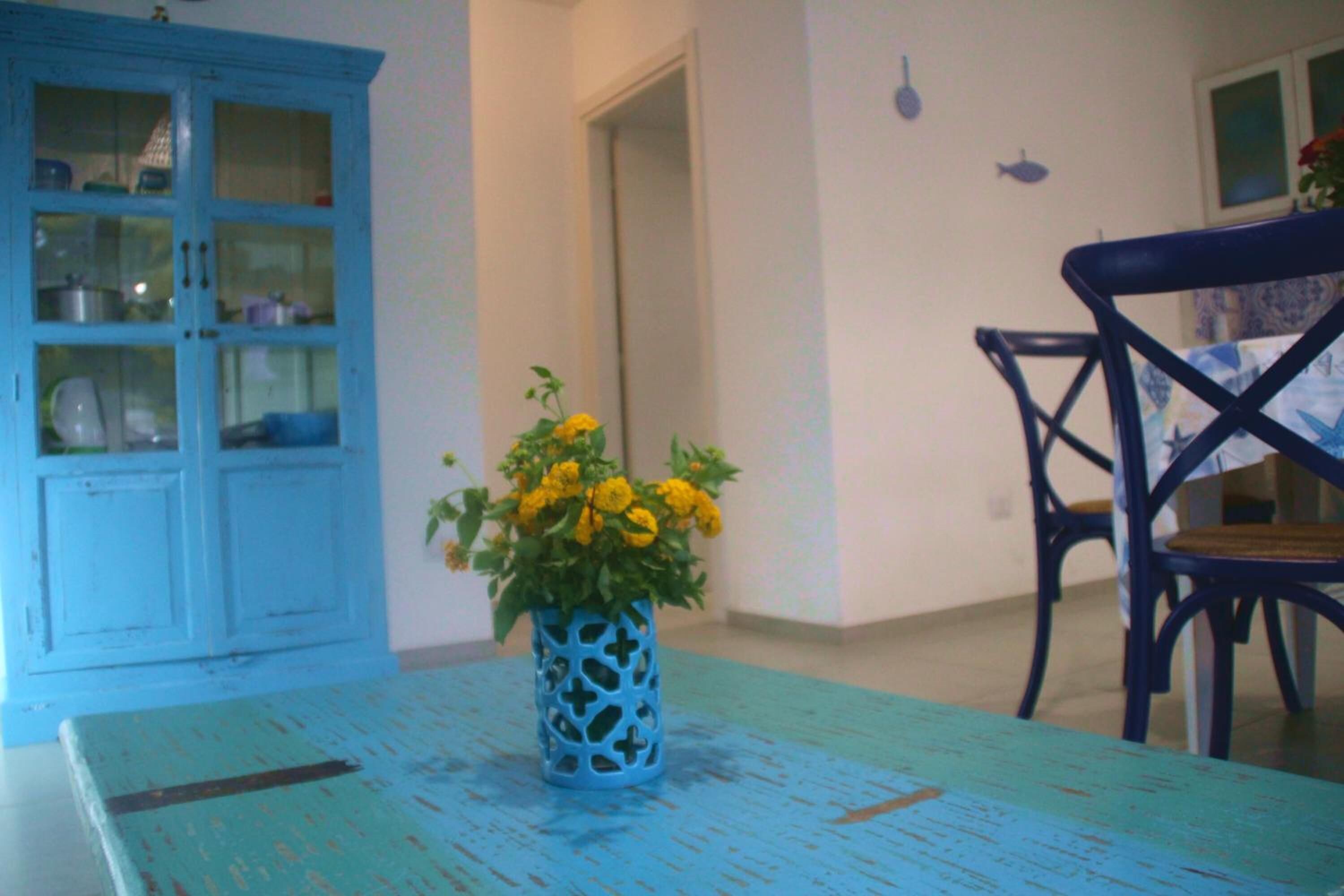 Uma sala de jantar com uma mesa e cadeiras azuis, um armário azul com portas de vidro e um vaso com flores amarelas.
