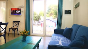 A living room with a blue sofa, a wooden table with a vase of flowers, a TV mounted on the wall, and a sliding glass door leading to an outdoor area with bicycles.