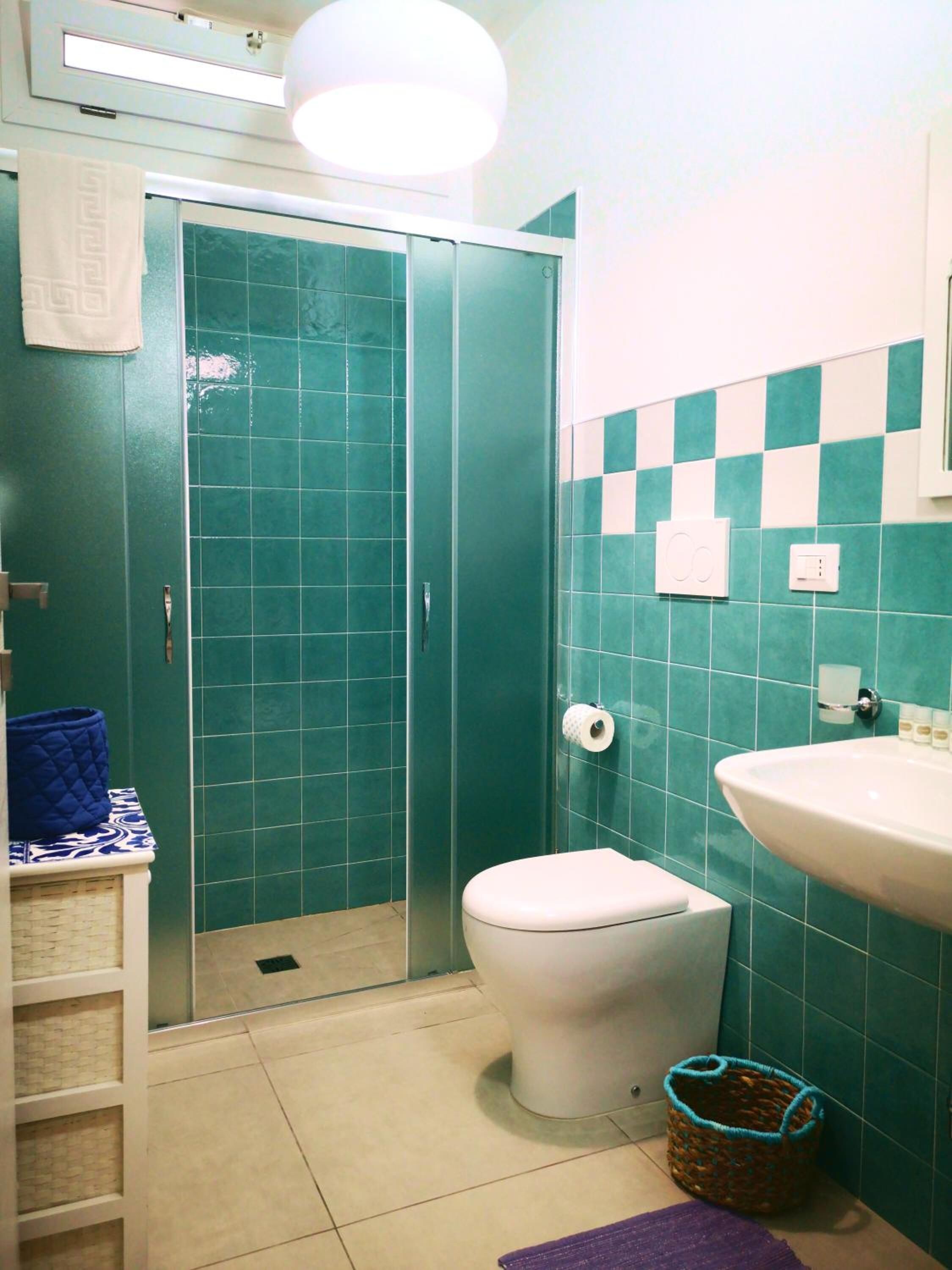 A bathroom with teal tiles, a white toilet, a sink, a shower, and a towel hanging on the wall.