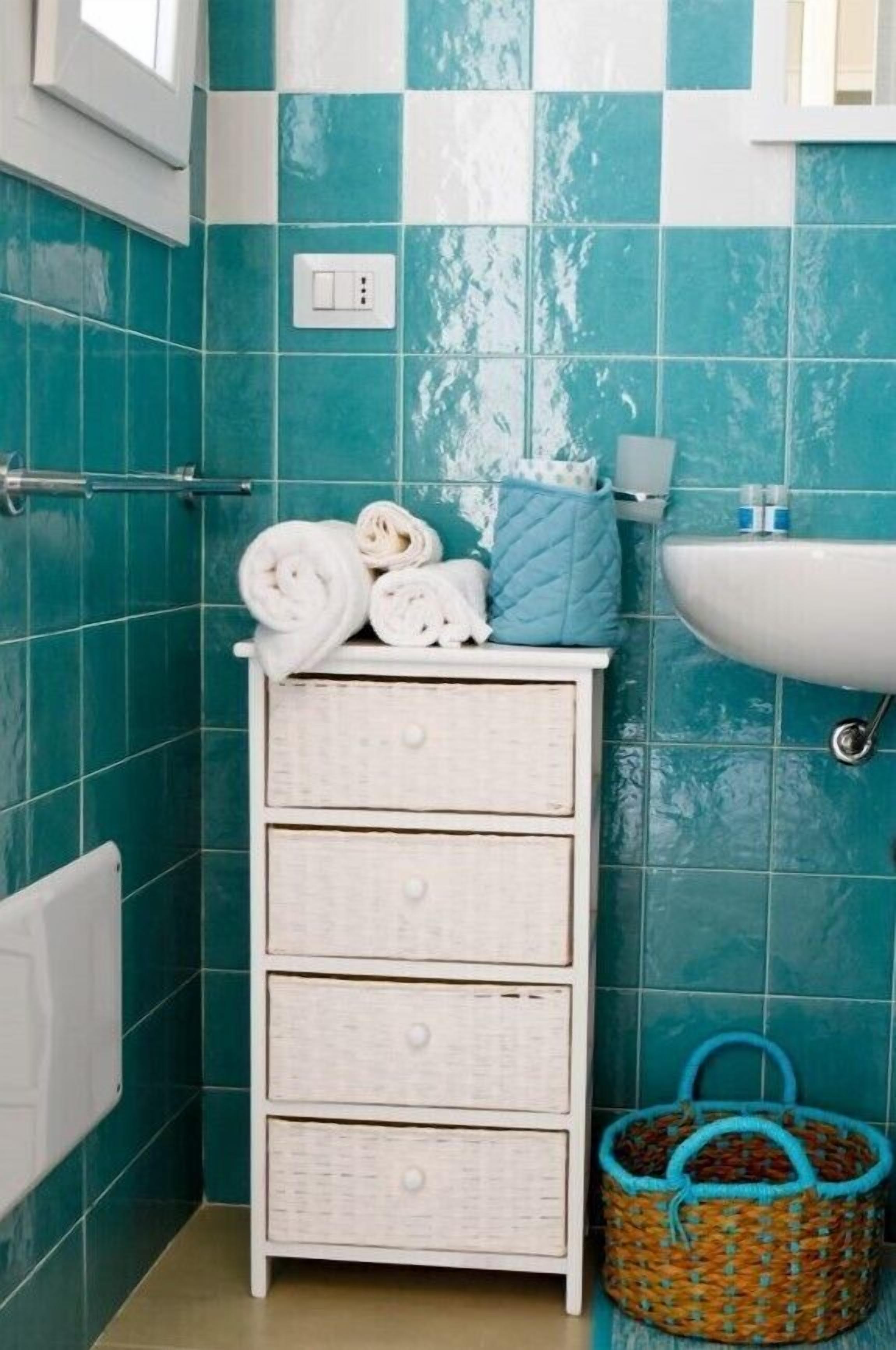 A bathroom with teal tiles, a white dresser with drawers, a white sink, and a wicker basket.