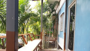 A wooden deck with a metal railing, surrounded by lush green foliage and a blue building with barred windows. - Nakhonyok Homestay (Nakhon Nayok)