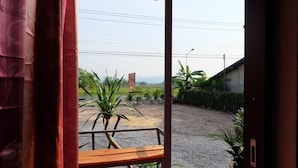 A view from a room with a wooden bench and a red curtain. Outside, there is a dirt ground, a few plants, and a building in the distance. - Nakhonyok Homestay (Nakhon Nayok)