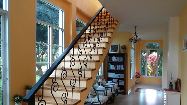 Una escalera con barandillas negras decorativas lleva a una zona de descanso con estantería, asientos y vistas al exterior, con vegetación.