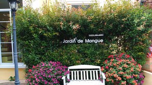 Un banco blanco frente a un letrero que dice "Jardin de Mangue", con plantas y flores en macetas alrededor.