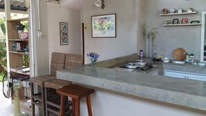 Una cocina con encimera de mármol, dos taburetes de madera, un ventilador, una estantería con libros y un cuadro enmarcado.