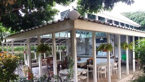 Un área de comedor al aire libre, cubierta, con mesas y sillas, rodeada de vegetación y un edificio al fondo.