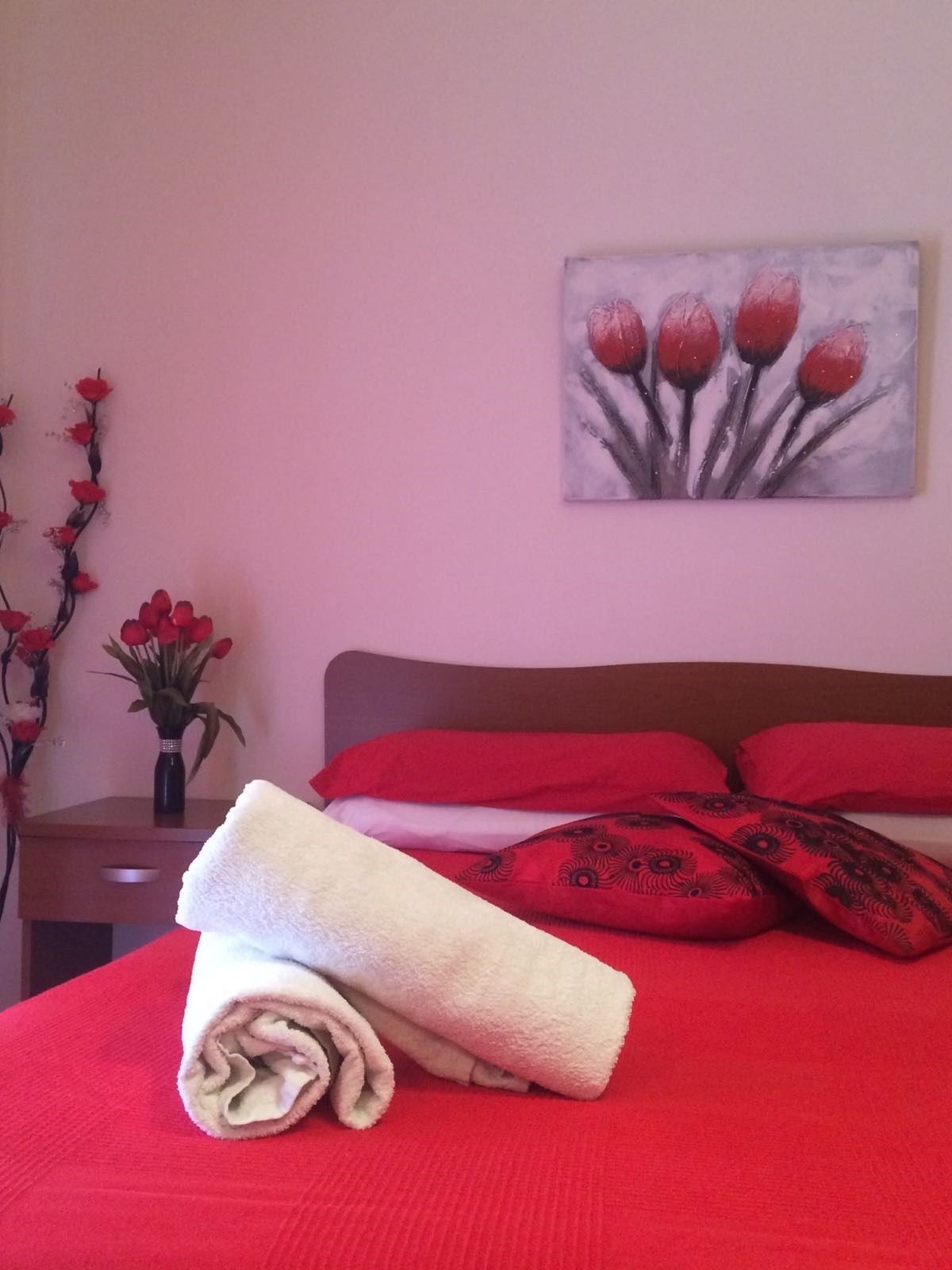 A neatly made bed with red bedding, a rolled white towel, and a painting of red tulips on the wall.