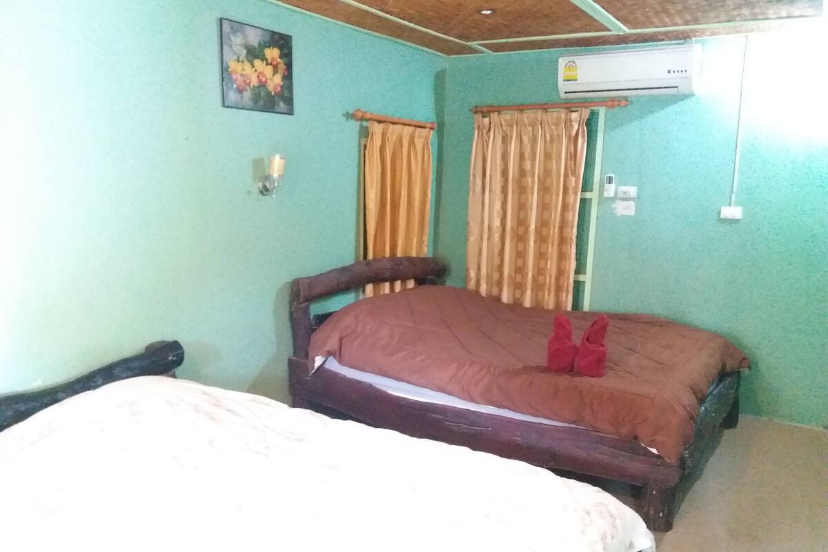 A single bed with a wooden frame and a brown mattress, a wall-mounted air conditioner, and a window with curtains.