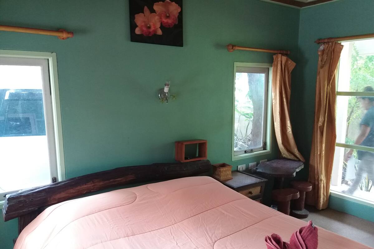 A room with a bed, a small table, a window with curtains, and a wall-mounted light fixture.