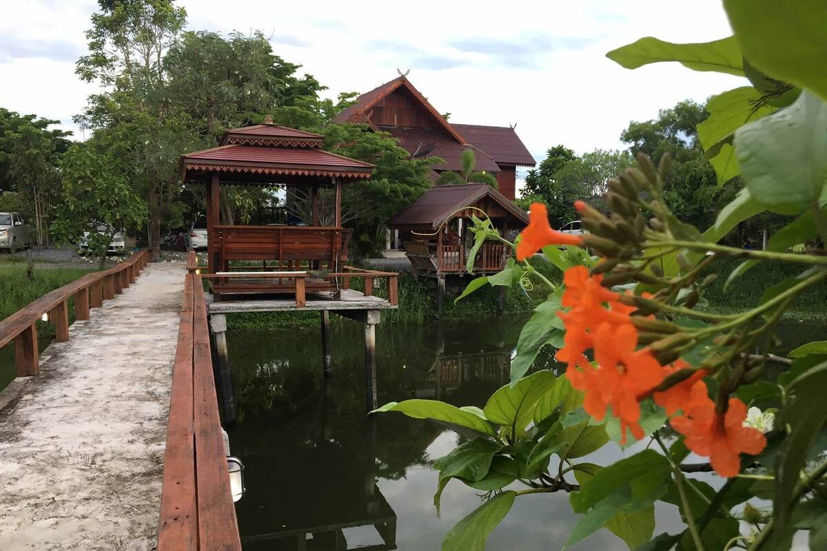 A wooden walkway leading to a traditional wooden structure over water, with a large house in the background.