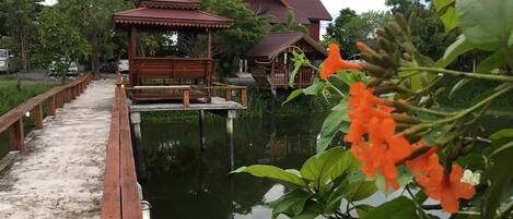 A wooden walkway leading to a traditional wooden structure over water, with a large house in the background.