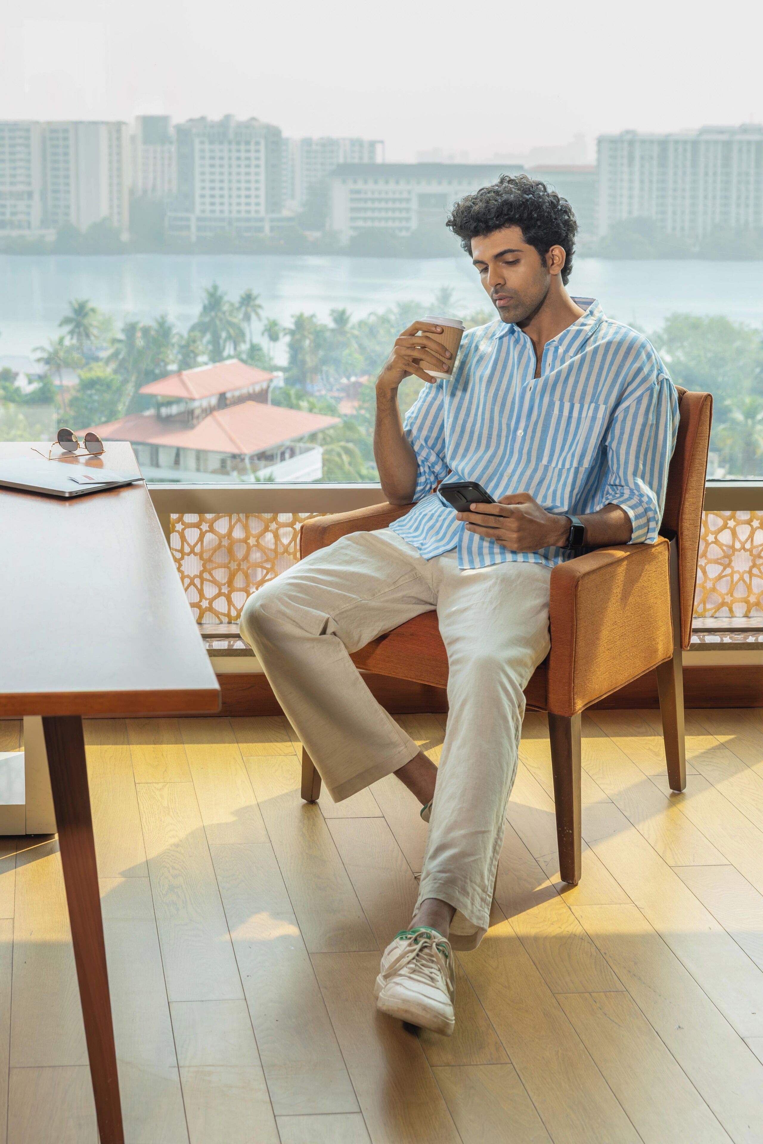 A person seated on a chair with a laptop and a cup on a table, overlooking a cityscape with buildings and palm trees.