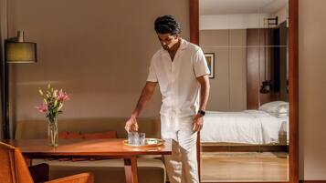 A hotel room with a bed, a dining table with chairs, a vase with flowers, and a bedside table with glasses and a bowl.