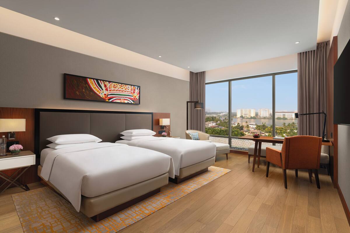 Ein Hotelzimmer mit zwei Betten, einem Schreibtisch, einem Sessel und einem großen Fenster mit Blick auf die Stadt. An der Wand hängt ein Gemälde.