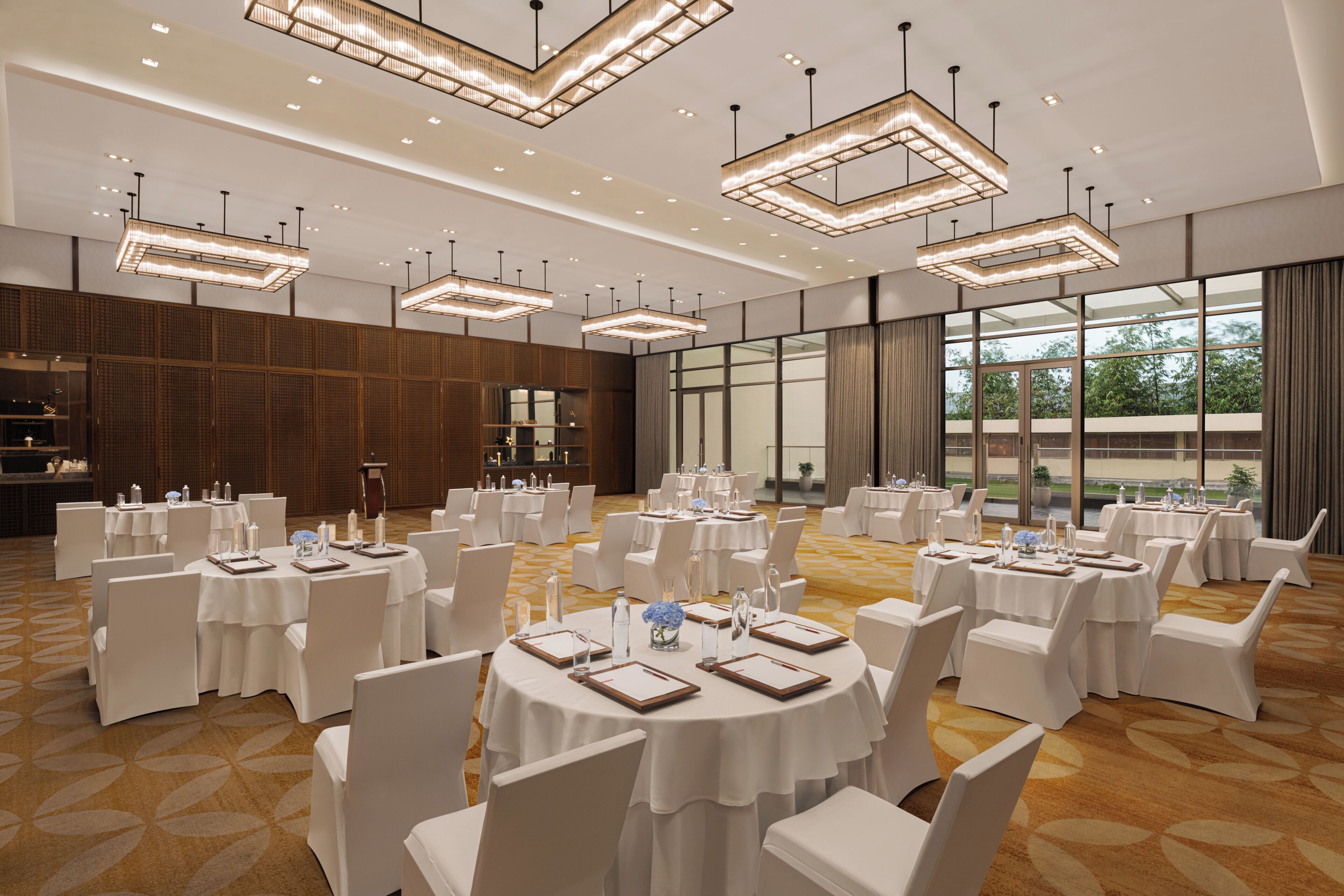 Un amplio salón de banquetes con mesas redondas, sillas blancas e iluminación colgante moderna.