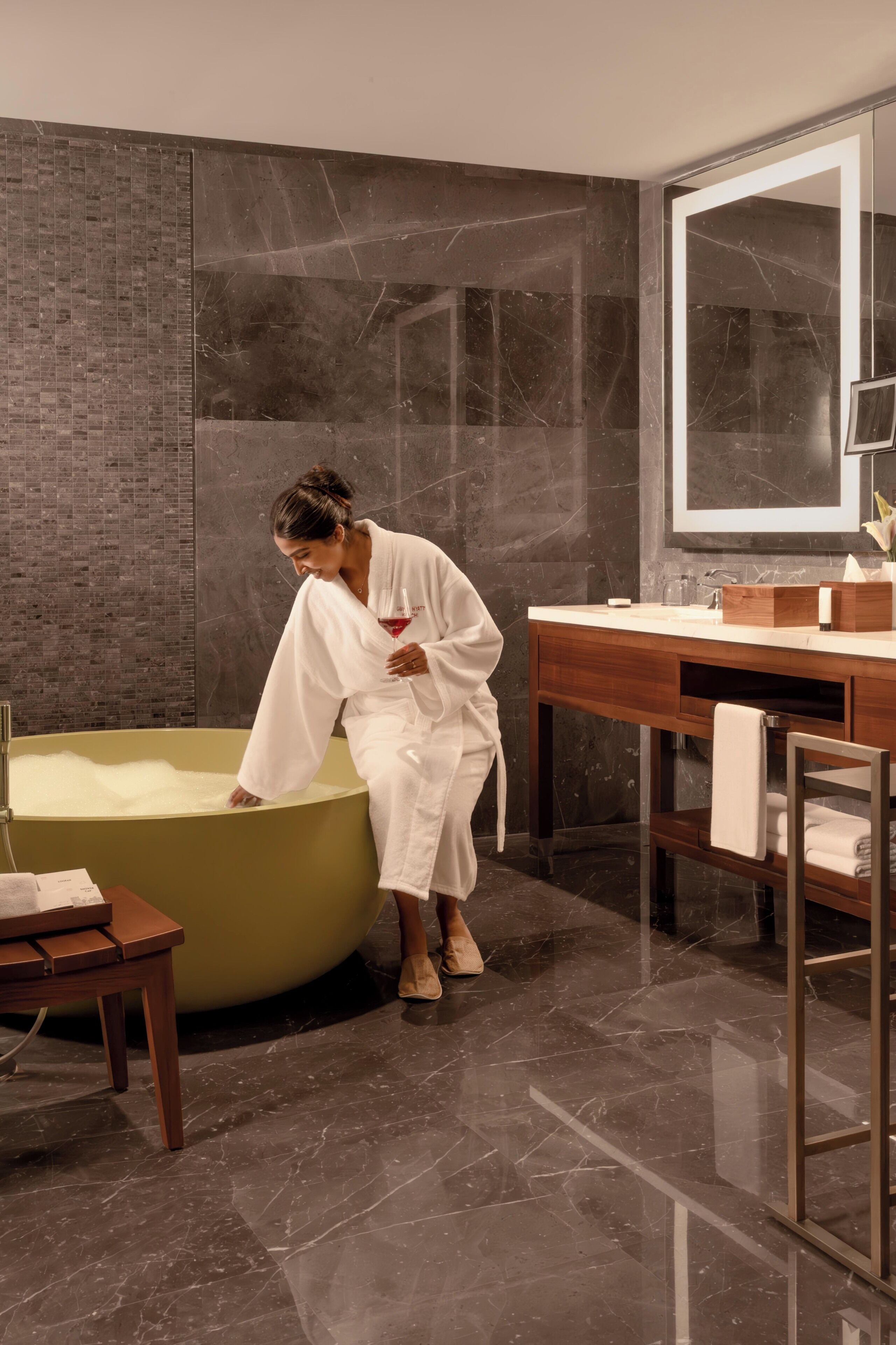 A person in a white bathrobe leaning into a large bathtub in a modern bathroom with dark marble walls and a wooden vanity.