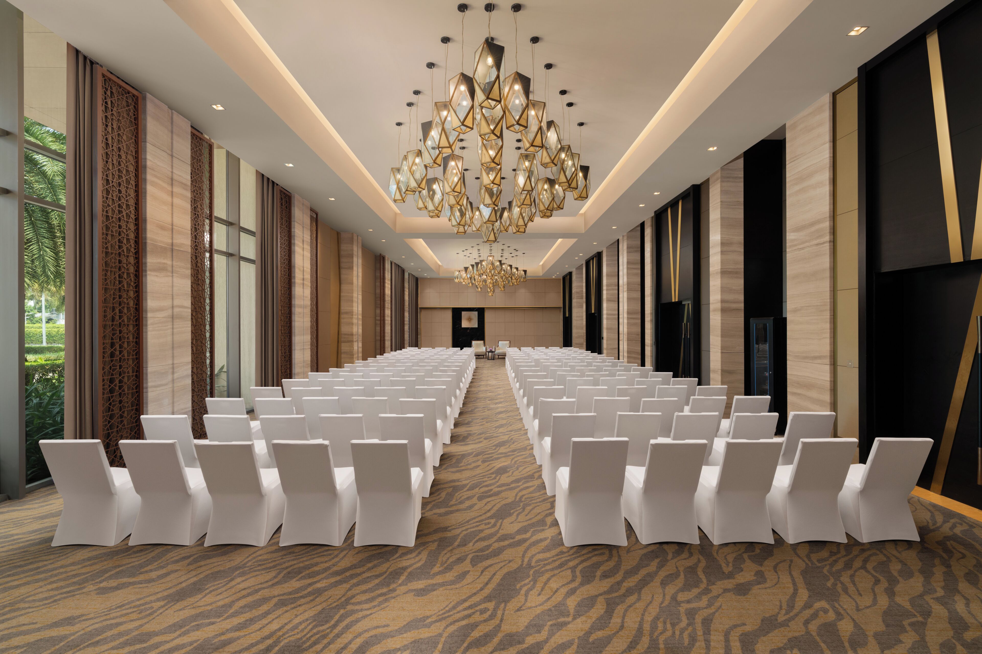Uma sala de conferências com fileiras de cadeiras brancas, um grande candelabro e um tapete estampado.